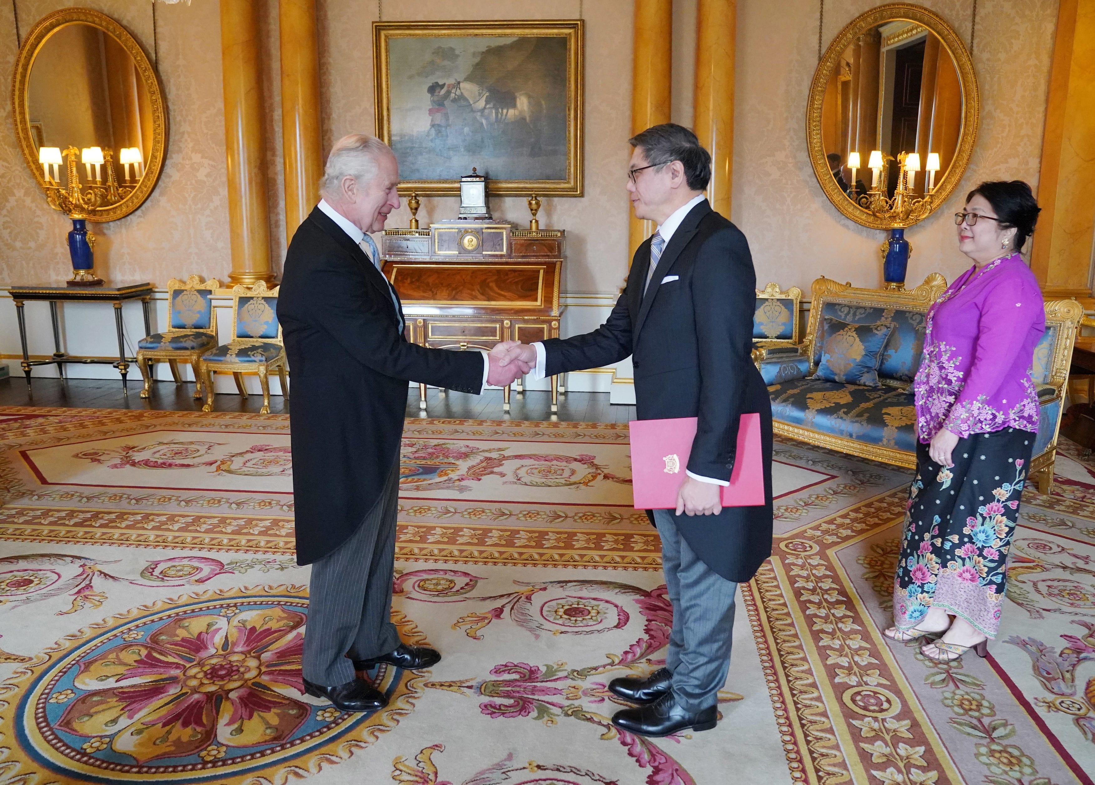 Two men in suits shaking hands, woman in purple and floral dress, ornate room.
